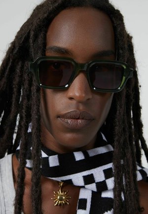 Lunettes de soleil en plastique vert avec une forme rectangulaire et des verres foncés. Le modèle porte une écharpe à carreaux noir et blanc et un pendentif soleil en or.