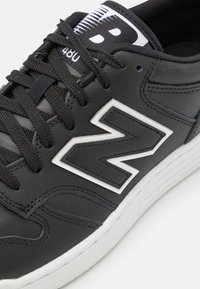 Baskets noires New Balance avec une semelle intermédiaire blanche, des lacets noirs et un grand logo "N" bordé de blanc sur le panneau latéral.