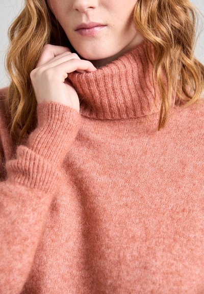 Knitted sweater in a soft, muted coral color. Features a ribbed turtleneck and wide cuffs with a fluffy texture, creating a cozy appearance.