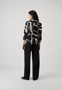 Blusa nera e crema con motivi geometrici, maniche a tre quarti e texture morbida, abbinata a pantaloni neri morbidi e scarpe a punta.