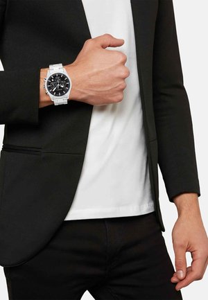 Sølv metalur med en sort urskive, der har chronograph-funktioner, på et håndled iført en sort jakke og en hvid T-shirt.