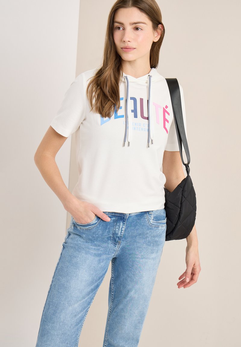 Jeune femme portant un sweat à capuche blanc à manches courtes avec un texte coloré, jean bleu, sac à bandoulière noir matelassé, debout devant un fond neutre.