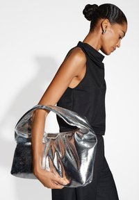 Borsa in argento metallico con una forma arrotondata e morbida. Presenta una maniglia larga e una superficie texturizzata, tenuta nella mano di una figura vestita di nero.