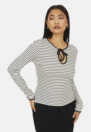 Vrouw met een zwart-wit horizontaal gestreepte top met lange mouwen en een keyhole-halslijn waarvan het uiteinde is vastgebonden met een zwarte strik, en zwarte broek.