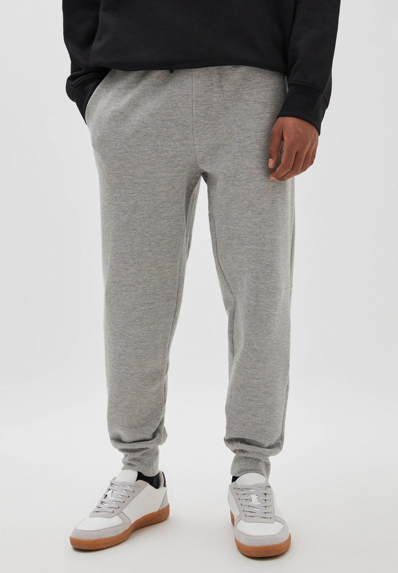 Personne portant un pantalon de survêtement gris clair avec des poignets élastiques et des baskets blanches à semelles en gomme, debout devant un fond uni.