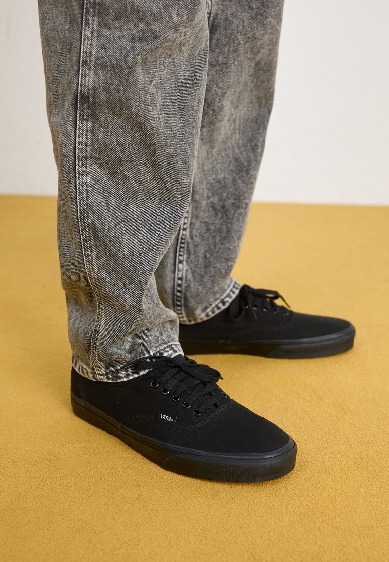 Personne portant des baskets Vans noires et un jean gris délavé debout sur un sol jaune moutarde.