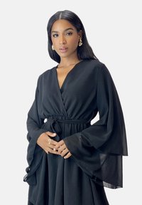 Zwarte wikkeljurk met lange, vloeiende mouwen, elastische taille en een geplooide rok. Zachte stof met een gladde textuur. Gouden ring aan de hand.