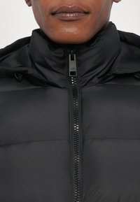 Doudoune noire avec un col haut, une fermeture éclair centrale avec logo, et une surface lisse et texturée. Design léger et rembourré.