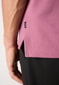 Rosa bomullspolo med sidsplit hems, med en liten svart "BOSS" etikett. Svarta byxor syns delvis i bakgrunden.