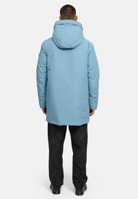 Garšs zils pūkainais mētelis ar kapuci, ar gludu tekstūru un nedaudz pagarinātu siluetu, apvienots ar tumšām biksēm un melno apavu.