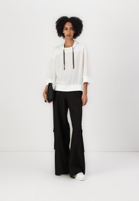 Weißer transparenter Hoodie mit schwarzen Kordeln, kombiniert mit lockeren, schwarzen weiten Hosen. Das Model hält eine schwarze Handtasche und trägt weiße Turnschuhe.
