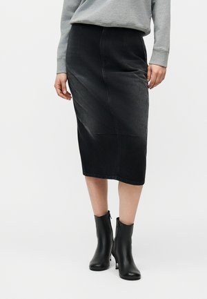 Personne portant une jupe noire jusqu'aux genoux, un sweat-shirt gris, et des bottines à talons noires, debout devant un fond blanc uni.