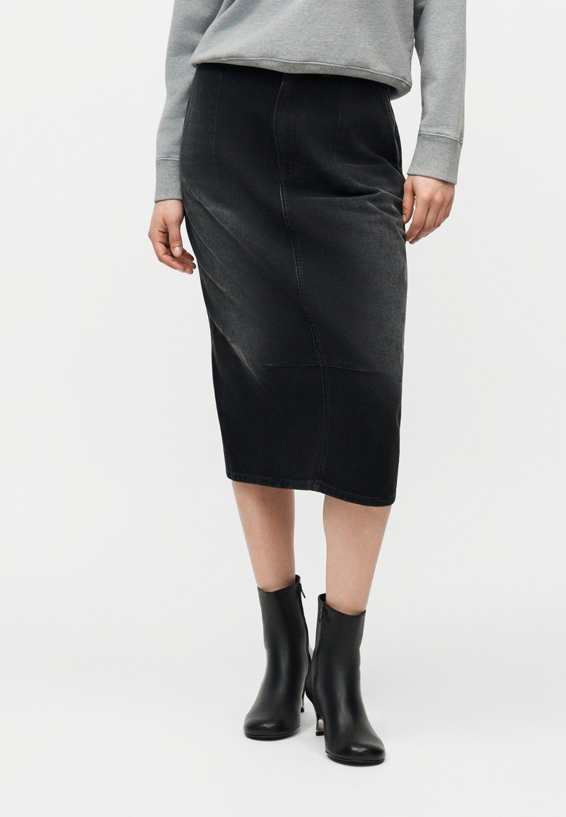 Άτομο που φοράει μαύρη φούστα μήκους μέχρι τα γόνατα, γκρι φούτερ και μαύρα μποτάκια με τακούνι, στέκεται μπροστά σε απλό λευκό φόντο.