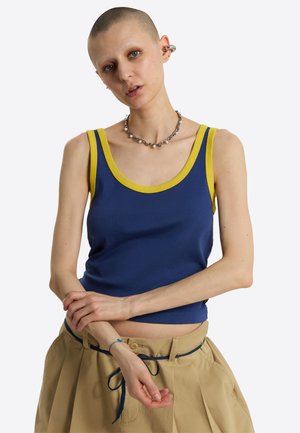Ung person med barberet hoved iført en blå tanktop med gul kant, beige bukser med blåt snøre bælte og sølv halskæde og øreringe.