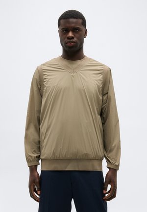 Homme portant un pull beige à manches longues avec des poignets élastiques et un pantalon bleu marine, debout devant un fond clair uni.