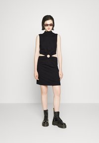 Vestido preto sem mangas com um decote alto, apresentando um detalhe torcido na cintura e um acento circular. Combinado com botas pretas de atacadores.