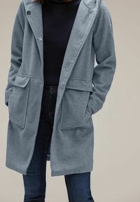 Ljusblå herringbone-jacka med huva, två framfickor och knappar. Bärs över en mörk topp och matchas med mörka jeans. Mjuk tygtextur.