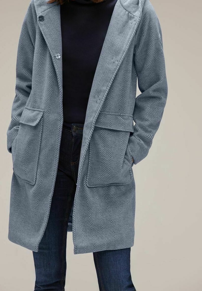 Ljusblå herringbone-jacka med huva, två framfickor och knappar. Bärs över en mörk topp och matchas med mörka jeans. Mjuk tygtextur.