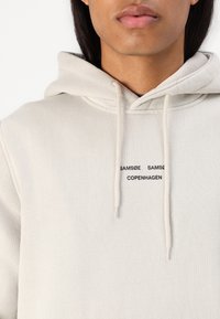 Il sudario grigio chiaro con cappuccio presenta una texture a coste, un cordino e una stampa del marchio: "SAMSE" e "COPENHAGEN" in nero sul davanti.