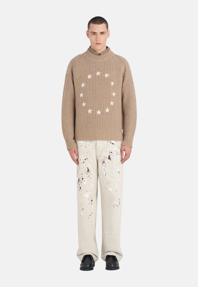 Pull en tricot beige avec un col montant et un motif d'étoiles blanches. Associé à un pantalon crème orné de taches de peinture colorées. Chaussures noires.