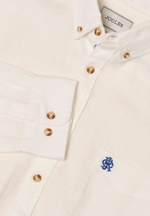 Hvid skjorte med knapper i brun farve, en blå broderet logo på lommen og et "Joules" mærke inden i kraven.