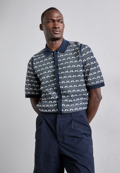 Polo en tricot à manches courtes de couleur bleu marine avec un motif géométrique blanc, doté d'un col uni et d'une fermeture à boutons, en tissu texturé.