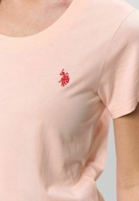 Svetloružové bavlnené tričko s okrúhlym výstrihom a krátkymi rukávmi, s červeným vyšitým logom na ľavej strane hrudníka.