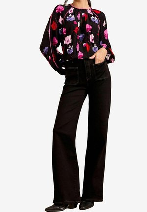 Vrouw draagt een zwarte blouse met lange mouwen en bloemenprint en een zwarte, hooggetailleerde broek met brede pijpen en voorzakken.