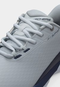 Zbliżenie na jasnoszary sneaker Skechers Slip-ins z teksturowaną tkaniną, wyściełanym językiem, białymi sznurówkami i granatowym detalem podeszwy.