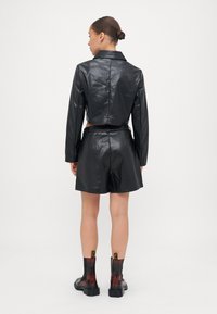 Veste courte en cuir noir avec un col, associée à un short en cuir noir. Le modèle porte des bottines transparentes noires à la cheville. Texture lisse.