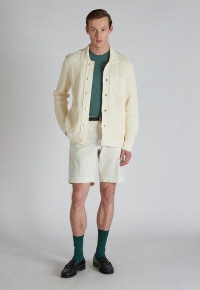 Cardigan a coste crema con grandi bottoni, t-shirt teal sotto, pantaloni corti chiari, calzini verdi e mocassini neri.