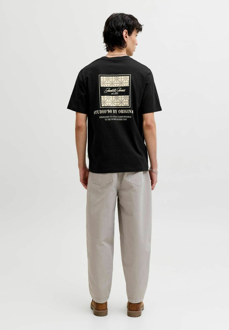 Homme portant un t-shirt noir à motifs avec texte et motifs beige, pantalon gris clair et chaussures marron, debout tourné de dos sur fond uni.