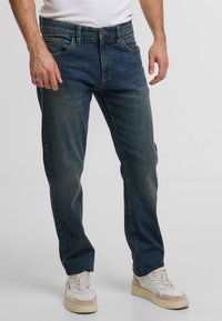 Jeans in denim blu con taglio dritto, cuciture visibili e tasche anteriori. Indossati con sneakers bianche e beige.