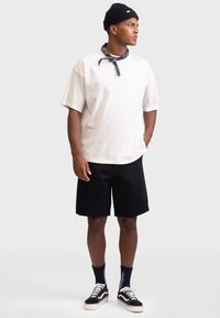 Cream short-sleeve t-shirt with a pocket, paired with black shorts. Accessories include a patterned bandana and black knit beanie. Black sneakers.