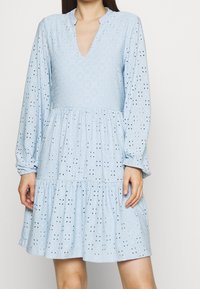 Abito azzurro chiaro con scollatura a V profonda, maniche lunghe, gonna a strati e dettagli in occhiello. Il tessuto testurizzato aggiunge interesse visivo.