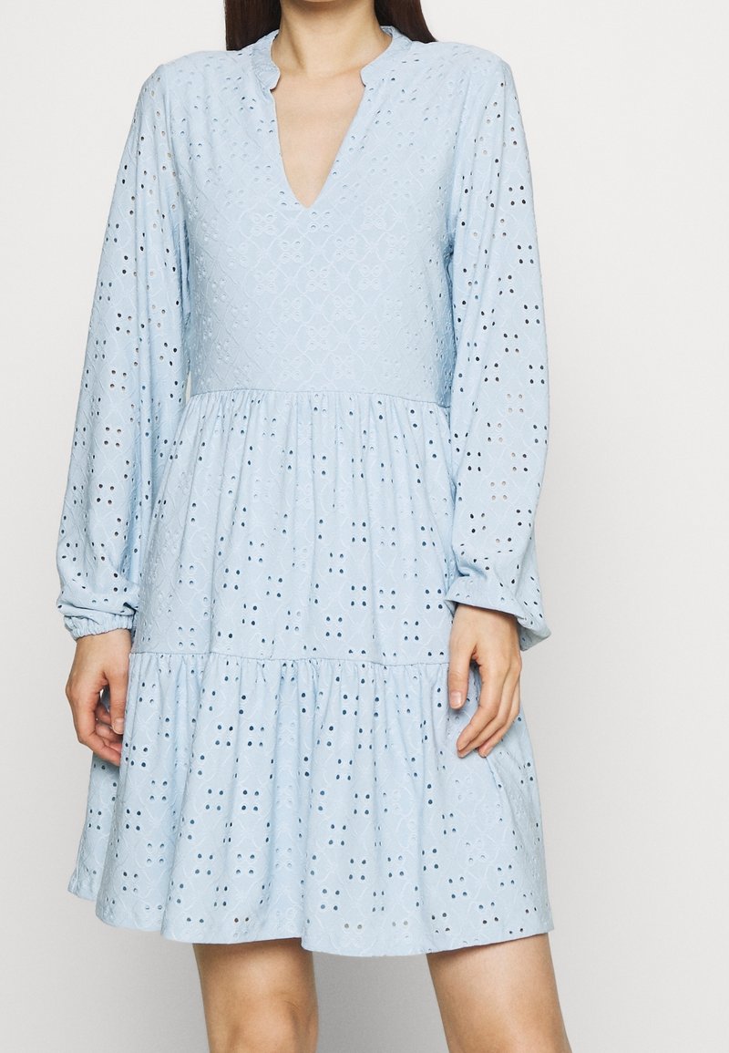 Abito azzurro chiaro con scollatura a V profonda, maniche lunghe, gonna a strati e dettagli in occhiello. Il tessuto testurizzato aggiunge interesse visivo.