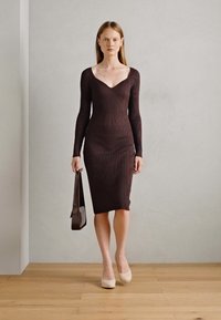 Abito lungo rosso bordeaux a maniche lunghe con texture a coste e scollo a V profondo. Silhouette aderente, lunghezza al ginocchio, abbinato a una borsa scura e décolleté chiari.