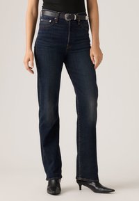 Σκούρα μπλε jeans με ίσια γραμμή από denim, με εμφανείς ραφές, μπροστινές τσέπες και μαύρη ζώνη. Συνδυάζονται με μαύρες μυτερές γόβες.