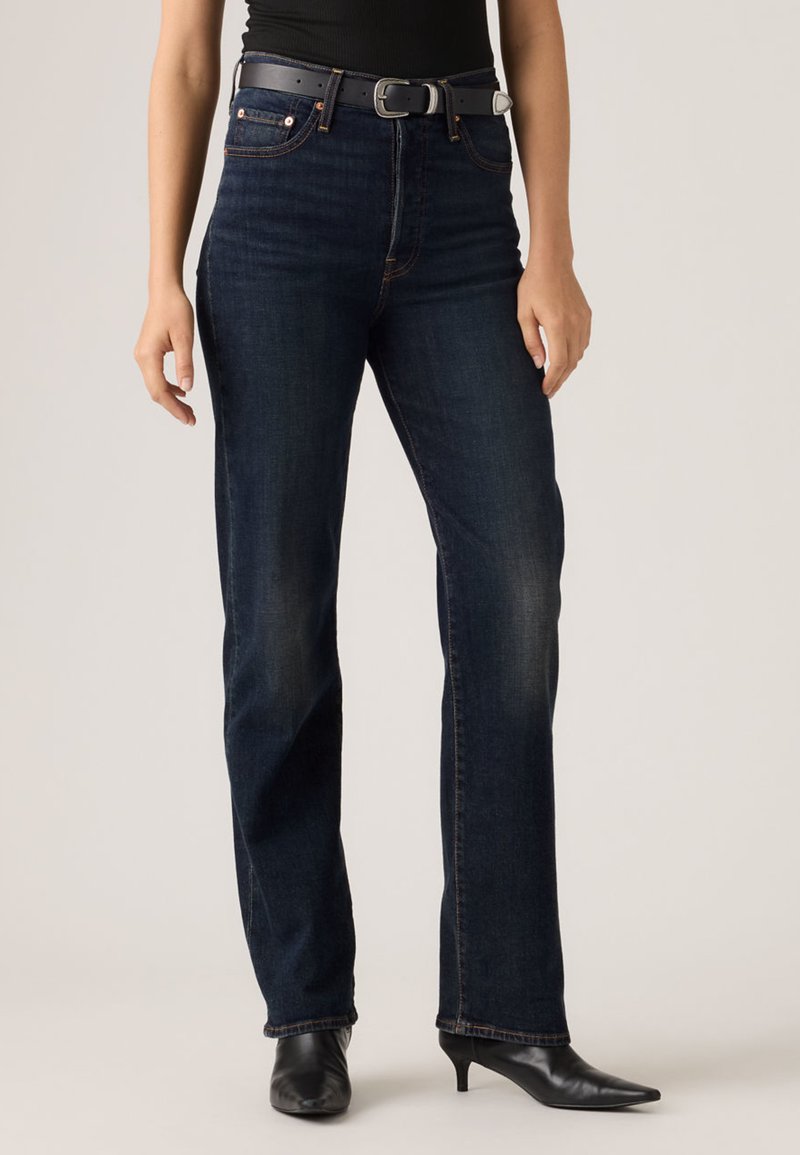 Σκούρα μπλε jeans με ίσια γραμμή από denim, με εμφανείς ραφές, μπροστινές τσέπες και μαύρη ζώνη. Συνδυάζονται με μαύρες μυτερές γόβες.