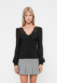Black top with sheer lace long sleeves, a deep V-neck, and fitted bodice; paired with a gray pleated skirt. Soft texture, intricate lace design.