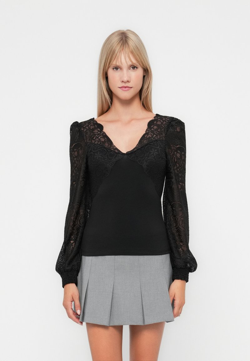 Black top with sheer lace long sleeves, a deep V-neck, and fitted bodice; paired with a gray pleated skirt. Soft texture, intricate lace design.