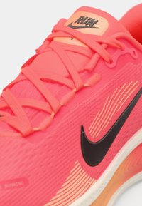 Chaussure de course Nike corail vif avec swoosh noir, lacets corail et étiquette "RUN" sur la languette, montrant un gros plan détaillé du côté et des lacets.