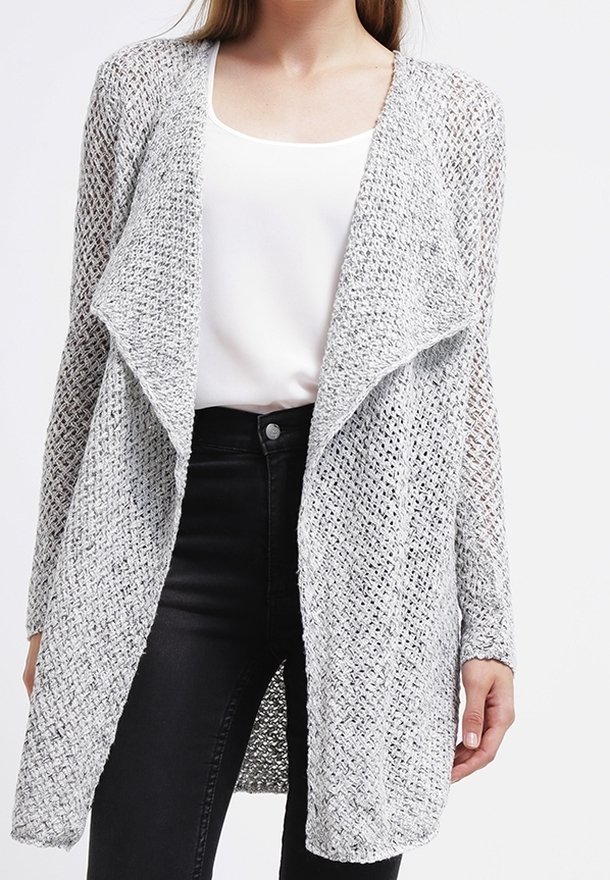 Femme portant un long cardigan gris clair à tricot ouvert sur un haut blanc et un jean noir taille haute, sur un fond uni.