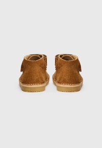 Botines de ante marrón claro con una textura suave, forma redondeada y suelas de goma beige en contraste. Presentan detalles de costura y lengüetas en la parte trasera.