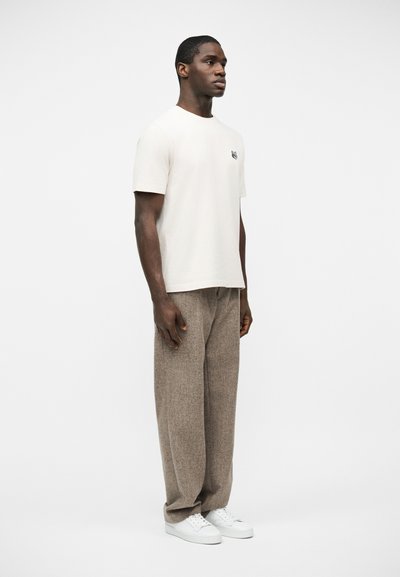 Άνδρας που στέκεται φορώντας άσπρο κοντομάνικο μπλουζάκι, φαρδιά καφέ υφασμάτινη παντελόνα και λευκά αθλητικά παπούτσια, στραμμένος ελαφρώς προς τα δεξιά σε απλό φόντο.