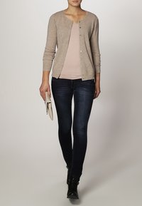 Beige stickad kofta med rund halsringning, knappar fram och långa ärmar, kombinerad med en ljusrosa topp och mörkblå smala jeans.