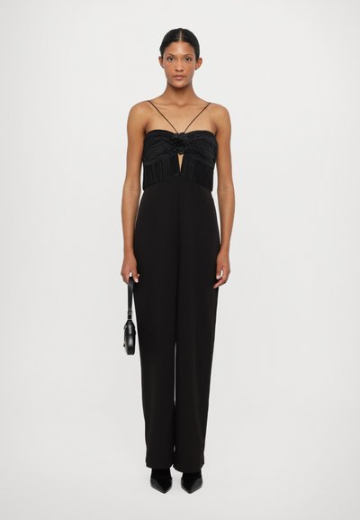 Zwarte jumpsuit met een geplooide buste, spaghettibandjes, een keyhole uitsnijding en wijde pijpen. Houdt een kleine zwarte handtas vast.