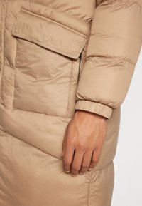 Beżowa puchowa kurtka o błyszczącej, gładkiej fakturze. Posiada dużą, skośną kieszeń na piersi oraz elastyczne mankiety dla lepszego dopasowania.