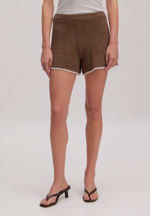 Vrouw draagt bruine gehaakte korte broek met witte rand en zwarte sandalen met hak, staand tegen een effen achtergrond.