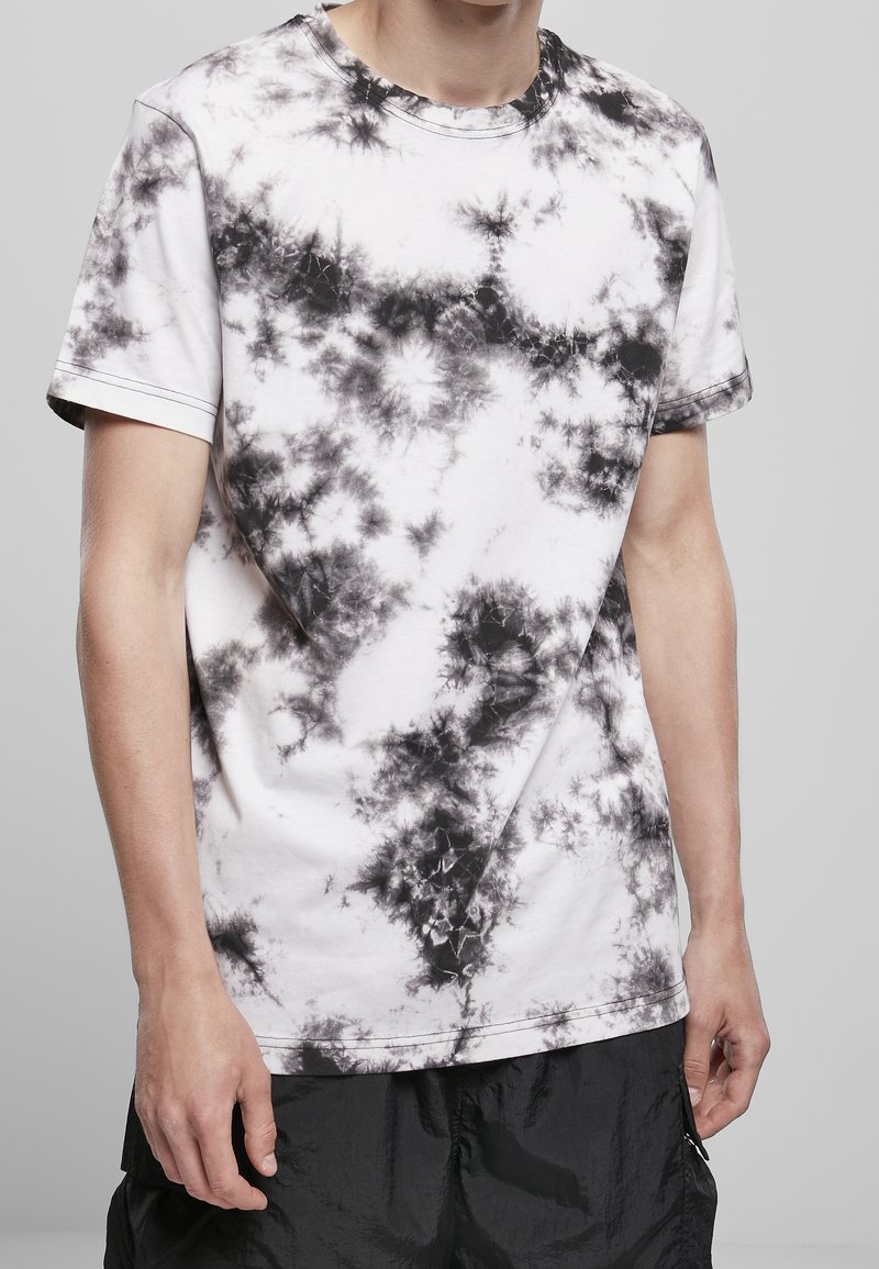 Homme portant un t-shirt blanc à manches courtes avec un motif tie-dye noir et un short noir ample, debout devant un fond uni.